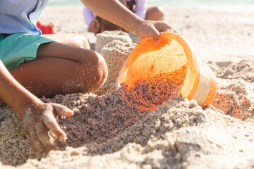 Midsection of boy collecting sand in bucket with sister playing at beach on sunny day