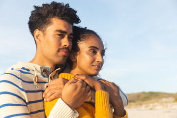 Thoughtful multiracial couple embracing while looking away at beach against sky on sunny day