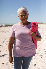 Portrait of smiling retired senior woman holding yoga mat with short white hair at beach