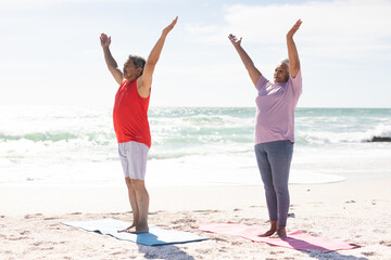 Full length of senior couple practicing yoga with arms raised at sunny beach against sky