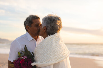 Newlywed senior multiracial couple kissing each other during wedding ceremony at beach