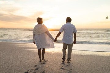 Naklejka premium Rear view of newlywed senior couple holding hands walking towards shore during sunset