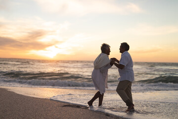 Full length of happy newlywed senior multiracial couple dancing at beach against sky during sunset