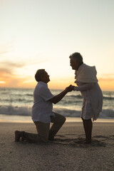 Fototapeta premium Side view of senior man proposing woman while kneeling on shore at beach during sunset