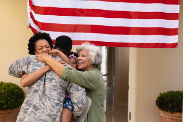Happy african american family embracing military soldier's return at entrance of house with flag