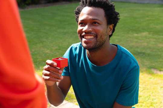 Smiling young african american man proposing girlfriend with engagement ring in garden