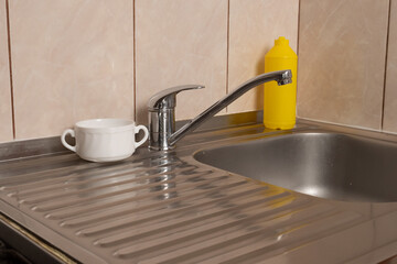 Metal kitchen sink with faucet, white cup and yellow dish soap bottle on draining board.