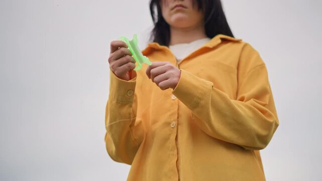 woman yellow jacket holding green toy, lowangle outdoor portrait under overcast sky, contemplative lone figure fidgets with plastic clip, textured fabric and button details, quiet cinematic mood