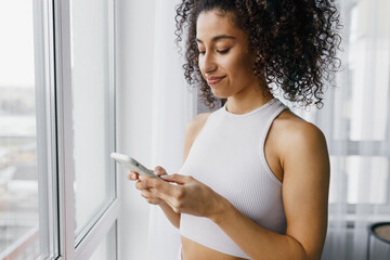 Closeup of happy attractive black girl in pink shirt texting love message to her boyfriend standing next to panoramic window of high-rise building, checking group chat with besties