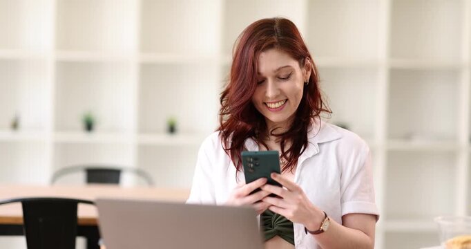 Focused busy caucasian businesswoman in a beige blazer sits at a modern office desk using her smartphone, checking important information or responding to a message with a concentrated expression
