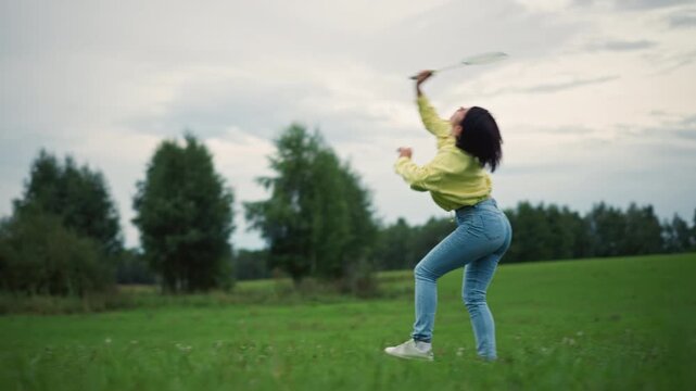 woman in yellow jacket moving in meadow, tossing small objects and reaching with controlled gestures slow fluid training sequence across green field under overcast sky, distant trees framing scene,