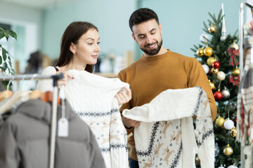 Obraz premium Joyful couple chooses cozy warm sweaters against the background of a Christmas tree in a decorated store. Man and a woman are preparing for the New Year holidays