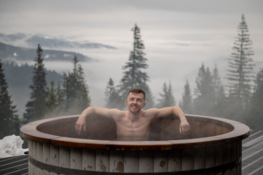 Tub. Man swims in hot bath while resting. Young male enjoying hot tub snowy forest outdoors. Winter holidays in snowy mountains. Guy relax in thermal spa in open air. Hot water treatment concept.
