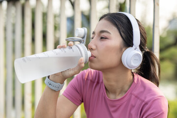Close-up of Asian woman in sportswear drinking water from bottle while wearing headphones, smartwatch after outdoor workout, symbolizing hydration, fitness recovery and healthy lifestyle.