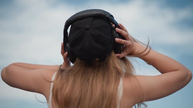 white woman listening to music outdoors, rear view with black cap and oversized headphones, long hair blowing in wind, hand adjusting headset, calm blue sky backdrop, streetstyle and DJ practice