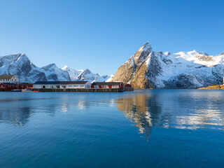 Lofoten Islands fishing village Reine with red rorbu cabins and a pier under a sunny sky in Norway. Crystal reflections on calm water, majestic mountain peaks, and traditional arctic summer scenery.