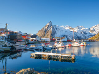 Lofoten Islands fishing village Reine with red rorbu cabins and boats at the pier under a sunny sky in Norway. Crystal reflections on calm water, majestic mountain peaks, and arctic summer vibe.