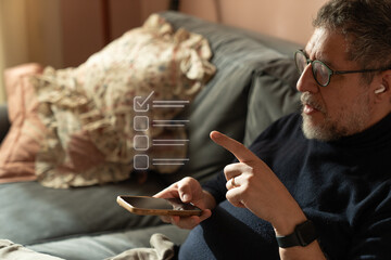 Middle aged man making digital decision at home using smartphone on sofa