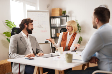 People in a modern office engage in a team meeting, sharing ideas and strategies. They sit around a table with papers and digital devices, focusing on teamwork and planning.