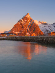 Vertical view of Reine and Hamnoy fishing villages at sunrise, pink glowing mountain peaks and traditional rorbu cabins. Crystal reflections on calm fjord water, Lofoten Islands, Norway.