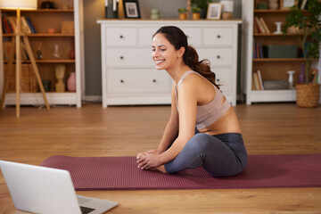 A woman performs a stretching exercise on a yoga mat in a well-lit living room. She appears focused and happy while following a workout on her laptop.