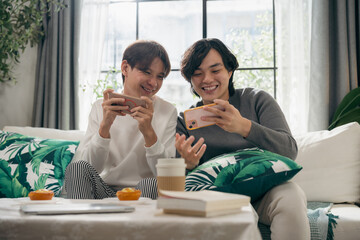 Happy Asian gay couple sitting together on a living room sofa, smiling while playing mobile games on a smartphone