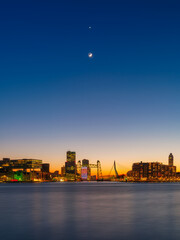 Vertical view of Rotterdam skyline at sunset. Modern skyscrapers and iconic bridges under a clear gradient sky. City center architecture, Erasmus Bridge, and urban twilight glow. Netherlands. 