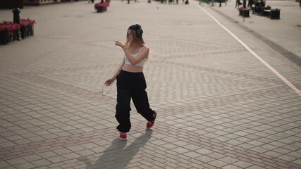street dancer moving across patterned pavement, female performer wearing cap, white tank top and baggy black pants with red sneakers freestyle arm and foot movements, relaxed confident energy, sunlit