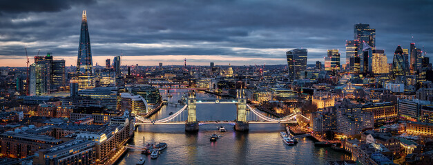 Compressed, panoramic aerial view of the illuminated Tower Bridge and city skyline of London, England, during twilight