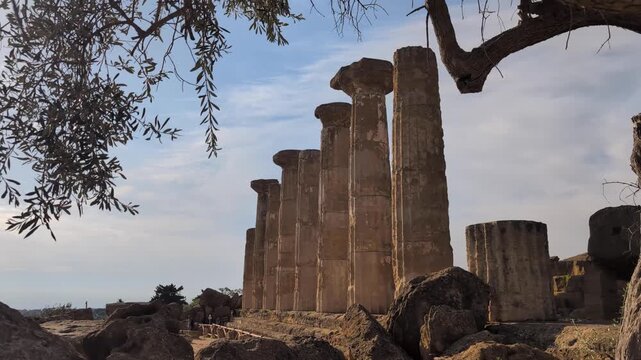 Ground-level view of ancient stone columns rising among rocks and olive branches at the Valley of the Temples, highlighting classical remains, erosion, and the timeless landscape of Agrigento.