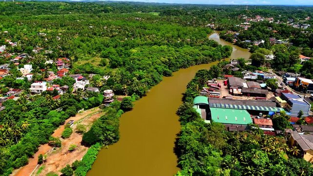 Aerial drone shot of the Nilwala River curving through Matara, with brown water, lush tropical vegetation and mixed riverside housing in bright daylight.