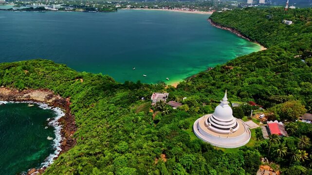 Aerial drone view of the Japanese Peace Pagoda in Rumassala overlooking a turquoise tropical bay, lush forested hills, rocky shorelines, and calm Indian Ocean waters in southern Sri Lanka.