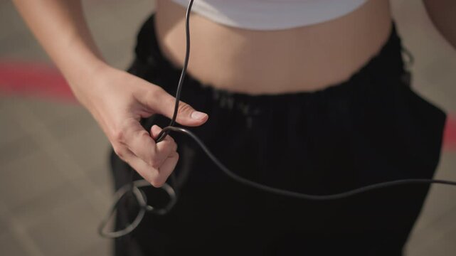 white female on street putting on headphones, plugging cable into smartphone and testing audio, fingers adjusting cord while midriff shows in crop top and black pants, hair catching sunlight, calm