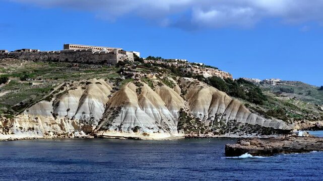 Wide low angle view tells the historic story of Fort Chambray atop dramatic striped clay cliffs in Gozo Malta during a bright spring day with deep blue sea water