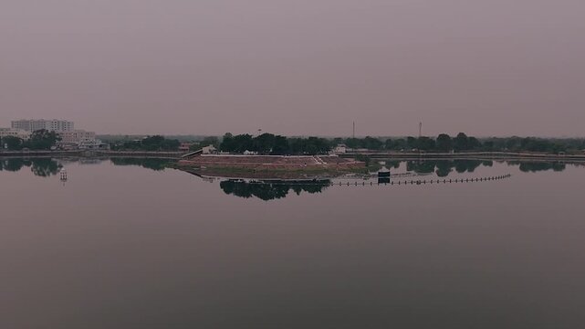Sharmishtha Lake Vadnagar Gujarat India showing wide calm water reflection, distant city edge and soft sky creating peaceful atmosphere for nature, travel, meditation and urban environment use.