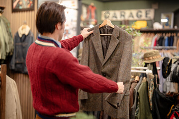 Fototapeta premium Hipster man on bargain hunting in fashionable vintage men's clothing store, found blazer and inspecting model and fabric quality of stylish item, pre owned goods shop selling selected fashion outfits