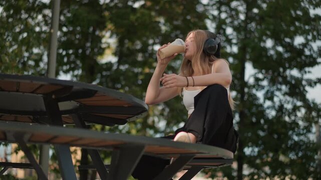 young caucasian woman on street bench sipping iced coffee while listening to headphones city buildings and skyline backdrop, playful dance gestures and relaxed poses, street performer energy turned