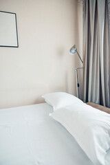 A clean, neatly made bed with white pillows and a soft blanket. A modern lamp stands beside the bed, and light curtains frame the window.