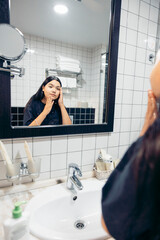 A young Hispanic woman with long dark hair applies skincare in a bathroom. She stands in front of a mirror, focusing on her facial care routine.