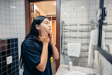 Young Asian woman with long black hair applying skincare in a modern bathroom. The scene emphasizes personal care and hygiene routines.