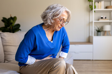 Senior woman suffering from abdominal pain while sitting on sofa at home