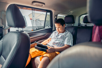 A young Hispanic boy sits in the back seat of a car, focused on a tablet. He wears a striped shirt and orange shorts, enjoying a trip.