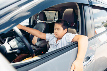 A young Hispanic boy sits in the driver's seat of a car, looking serious while resting his arm on the window. The interior is modern and spacious.
