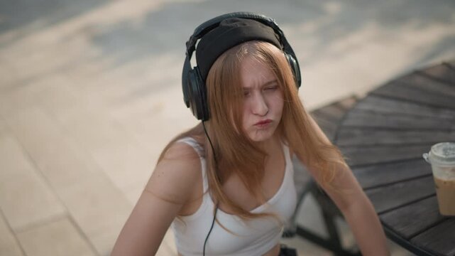 caucasian young woman dancing street cafe, wearing black beanie and overear headphones, white crop top and shorts, long red hair moving with rhythm, iced coffee on wooden round table, sunlight