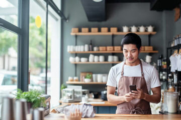 Fototapeta premium Young Asian Male Cafe Owner Using Smartphone at Counter