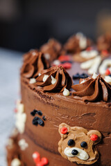 A rich layered chocolate cake with smooth buttercream, decorated with adorable teddy bear faces, piped swirls, and heart details, creating a playful and festive dessert centerpiece