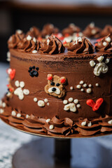 A rich layered chocolate cake with smooth buttercream, decorated with adorable teddy bear faces, piped swirls, and heart details, creating a playful and festive dessert centerpiece