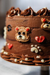 A rich layered chocolate cake with smooth buttercream, decorated with adorable teddy bear faces, piped swirls, and heart details, creating a playful and festive dessert centerpiece