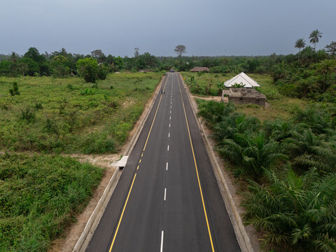 Aerial view of a straight black tar road cutting through lush green vegetation, with buildings on either side under a cloudy sky, Okobe, Rivers State, Nigeria.