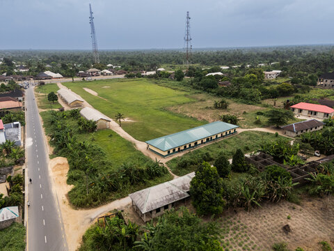 Aerial view of buildings with corrugated roofs and a green field under a cloudy sky, framed by lush trees, Okobe, Rivers State, Nigeria.